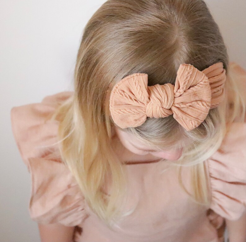May include: A light brown headband with a large bow tied in the center. The headband is made of a soft, textured fabric.