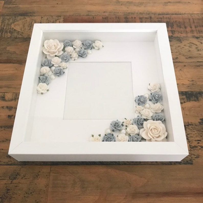 Puede incluir: Un marco de fotos blanco con un paspart&uacute; blanco y un fondo blanco. El marco est&aacute; decorado con flores de papel blancas y grises.