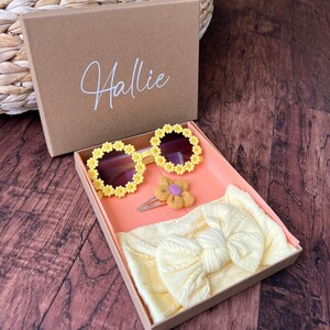 May include: A brown gift box with the name "Hallie" on the lid. The box contains a yellow headband, a pair of yellow sunglasses with flower accents, and a yellow flower hair clip.