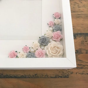 Puede incluir: Un marco de fotos blanco con un dise&ntilde;o floral decorativo. Las flores est&aacute;n hechas de papel y son de tonos rosa, gris y blanco. Las flores est&aacute;n dispuestas en cascada alrededor del borde del marco.