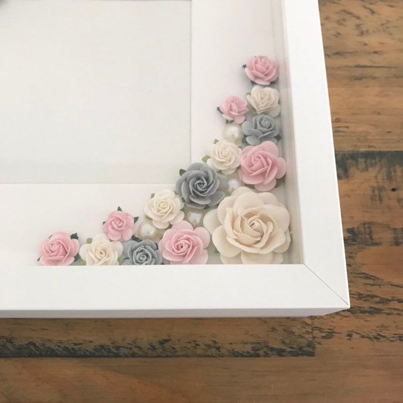 Puede incluir: Un marco de fotos blanco con un dise&ntilde;o floral decorativo. Las flores est&aacute;n hechas de papel y son de tonos rosa, gris y blanco. Las flores est&aacute;n dispuestas en cascada alrededor del borde del marco.