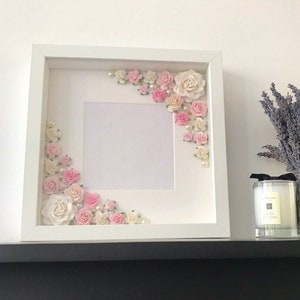 Puede incluir: Marco de fotos blanco con un paspart&uacute; blanco y un fondo blanco. El marco est&aacute; decorado con flores rosas y blancas y perlas.