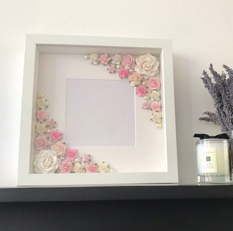 Puede incluir: Marco de fotos blanco con un paspart&uacute; blanco y un fondo blanco. El marco est&aacute; decorado con flores rosas y blancas y perlas.