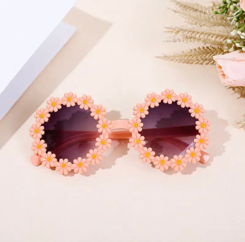 May include: Pink round sunglasses with a daisy flower design around the frames. The sunglasses have dark lenses.