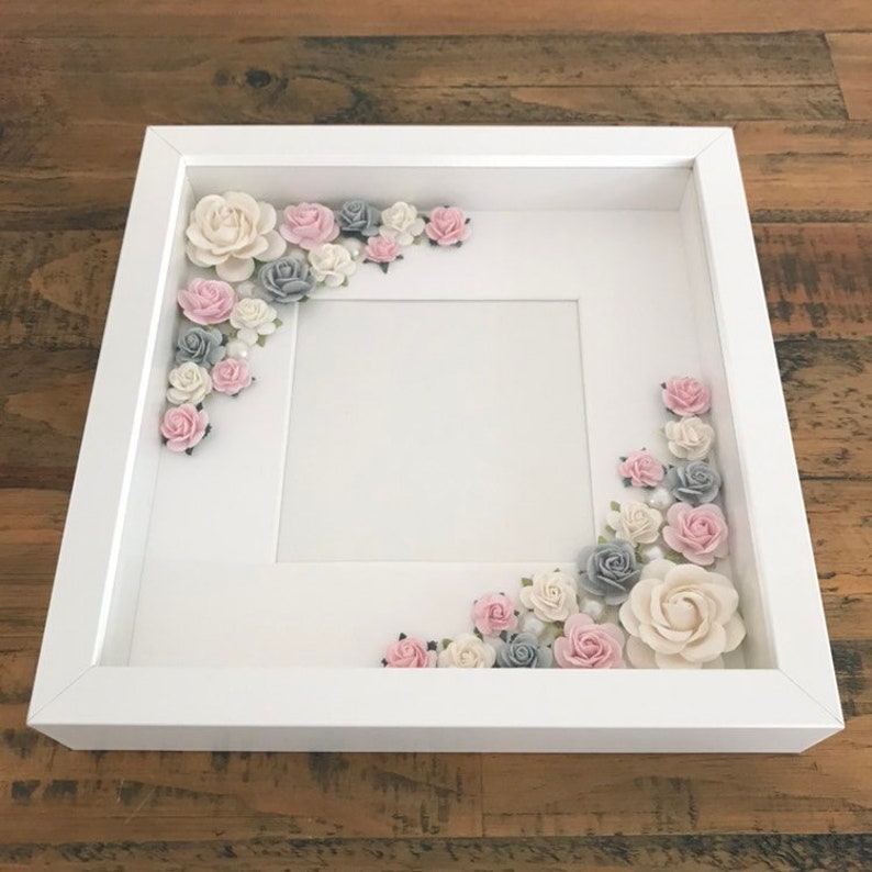 Puede incluir: Un marco de fotos blanco con un paspart&uacute; blanco y un fondo blanco. El marco est&aacute; decorado con flores de papel rosa, blanco y gris.