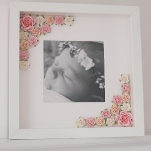 Puede incluir: Un marco de fotos blanco con una foto en blanco y negro de un beb&eacute; durmiendo. El marco est&aacute; decorado con flores de papel rosas y blancas.