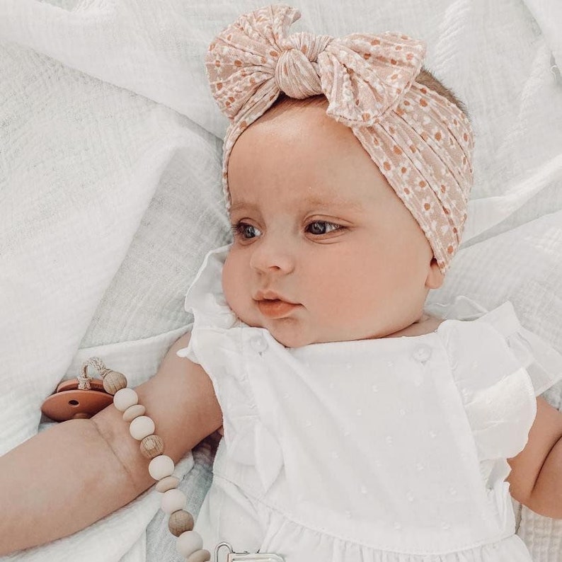 May include: A baby wearing a white dress with a pink floral headband. The baby is lying on a white blanket and has a wooden beaded pacifier clip.