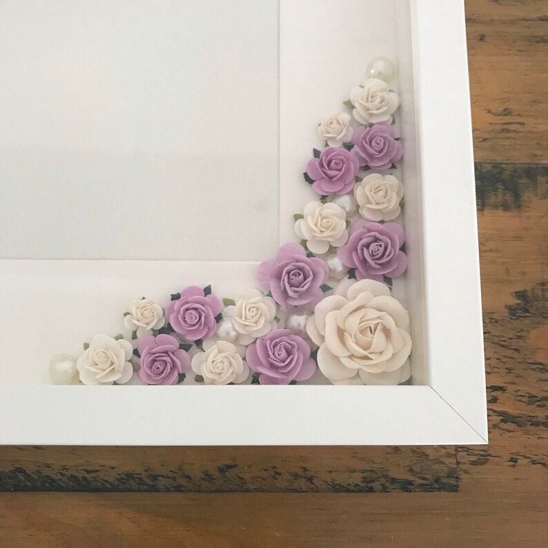 Puede incluir: Un marco de fotos blanco con una esquina llena de flores de papel moradas y blancas y detalles de perlas.