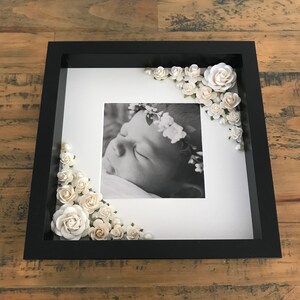 Puede incluir: Un marco de fotos negro con un paspart&uacute; blanco y una foto en blanco y negro de un beb&eacute; durmiendo que lleva una corona de flores. El marco est&aacute; decorado con flores de papel blanco y perlas.