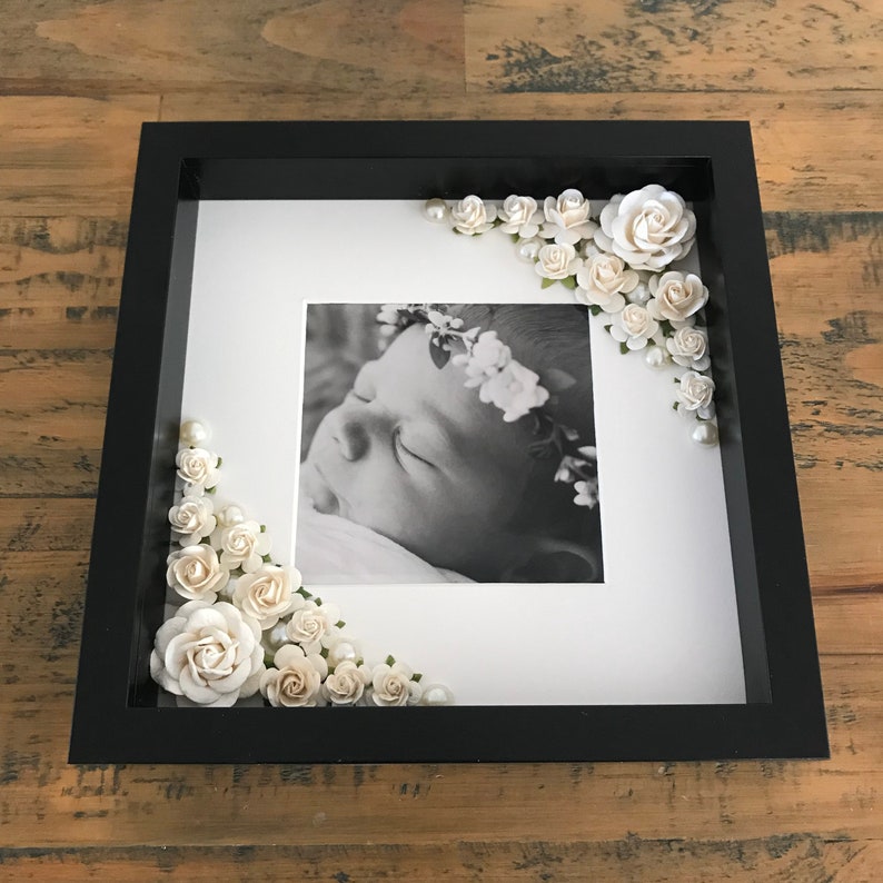 Puede incluir: Un marco de fotos negro con un paspart&uacute; blanco y una foto en blanco y negro de un beb&eacute; durmiendo que lleva una corona de flores. El marco est&aacute; decorado con flores de papel blanco y perlas.