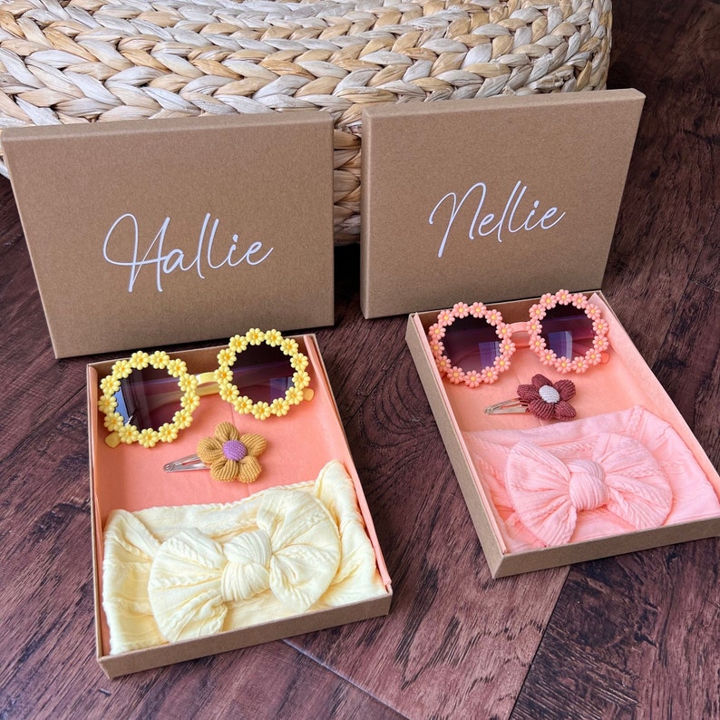 May include: Two brown gift boxes with lids open, each containing a headband, a flower hair clip, and a pair of yellow daisy-shaped sunglasses. The boxes are labeled "Hallie" and "Nellie".