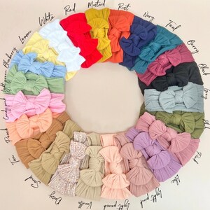 May include: A variety of colors of fabric headbands with a knotted bow. The colors include: blueberry, lemon, white, red, mustard, rust, navy, teal, berry, black, grey, tan, peach, candy pink, light green, lilac, light pink, light peach, vanilla, daisy, sand.