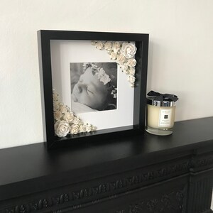 Puede incluir: Un marco de fotos negro con un paspart&uacute; blanco y una foto en blanco y negro de un beb&eacute; durmiendo. El marco est&aacute; decorado con flores blancas y perlas.