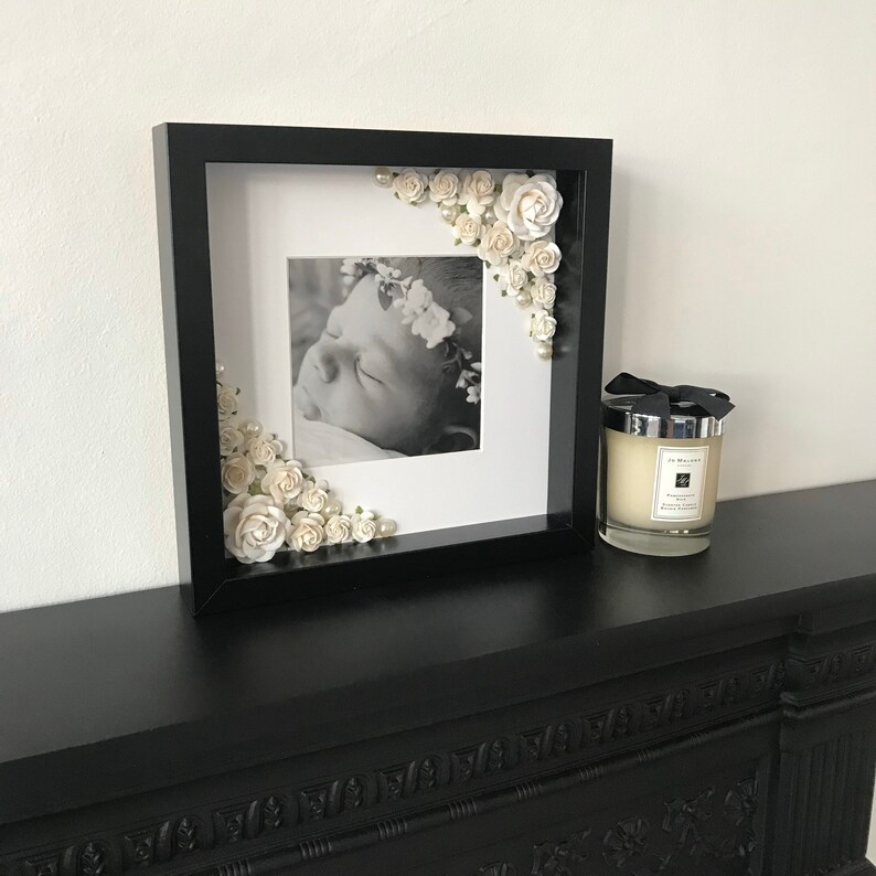 Puede incluir: Un marco de fotos negro con un paspart&uacute; blanco y una foto en blanco y negro de un beb&eacute; durmiendo. El marco est&aacute; decorado con flores blancas y perlas.