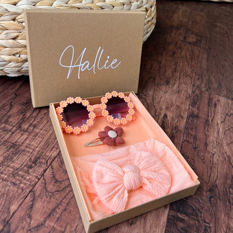 May include: A gift box with the name "Hallie" on the lid. The box contains a pair of peach-colored sunglasses with flower accents, a peach-colored headband with a bow, and a peach-colored flower hair clip.