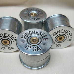 May include: Four silver shotgun shell caps with black lettering that reads "Winchester 12 GA".