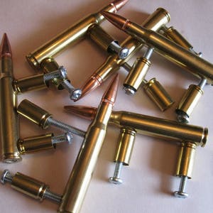 May include: A collection of brass bullet casings repurposed as hardware, featuring copper-coloured tips and silver-toned metal screws protruding from the sides. The metallic components are arranged in a scattered pile, showcasing the cylindrical brass bodies.
