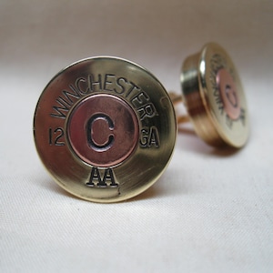 May include: A pair of gold cufflinks made from repurposed 12 gauge shotgun shells. The shells are engraved with "WINCHESTER" and "AA" and have a copper center with the letter "C" engraved on it.