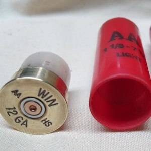 May include: Close-up of shotgun shells. One shell is assembled with a brass base and a clear top. The base is stamped with "AA", "12 GA", "WIN", and "HS". A red shell casing is also visible, with "AA" and "LIGHT" printed on it.
