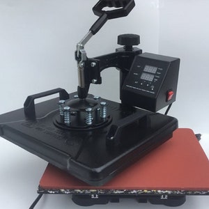 May include: Black heat press machine with a digital display and a red button. The machine has a round base with a spring-loaded clamp and a lever to lower the press. The machine is sitting on a red heat-resistant mat.