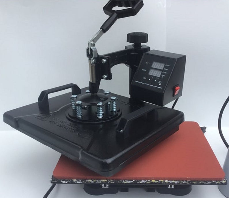 May include: Black heat press machine with a digital display and a red button. The machine has a round base with a spring-loaded clamp and a lever to lower the press. The machine is sitting on a red heat-resistant mat.
