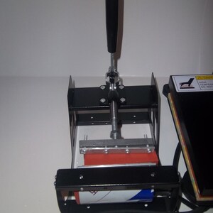 May include: A black heat press with a red heat plate and a lever handle. The heat press is used for transferring designs onto fabric.