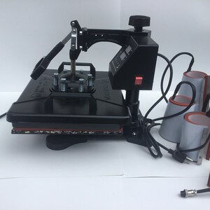 May include: Black heat press machine with a digital timer and a red power button. The machine has a flat surface for pressing items and comes with multiple attachments for different shapes, including mugs and hats.