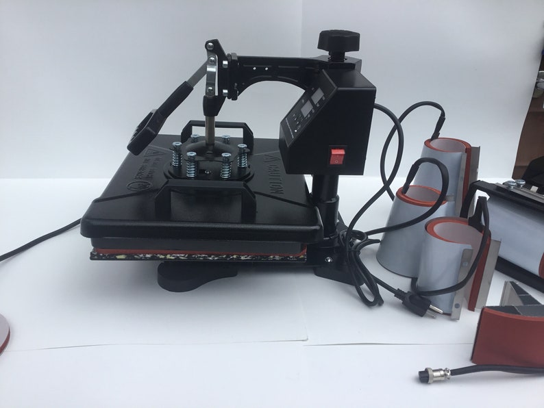 May include: Black heat press machine with a digital timer and a red power button. The machine has a flat surface for pressing items and comes with multiple attachments for different shapes, including mugs and hats.