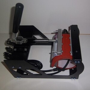 May include: Black mug press with a red heating element and a lever handle. The press is on a white surface.