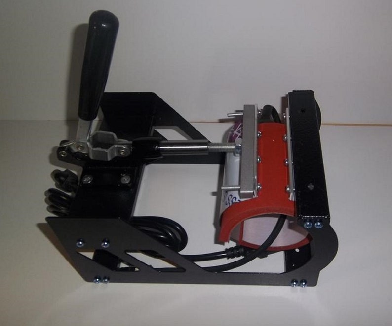 May include: Black mug press with a red heating element and a lever handle. The press is on a white surface.