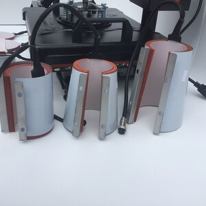 May include: Four white ceramic mug press attachments with red silicone and silver metal accents. The attachments are used for heat pressing designs onto mugs.
