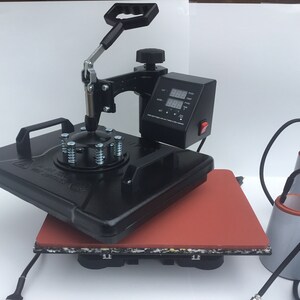 May include: Black heat press machine with a digital timer and temperature control. The machine is on a red heat-resistant mat. Three additional heat press attachments are included.