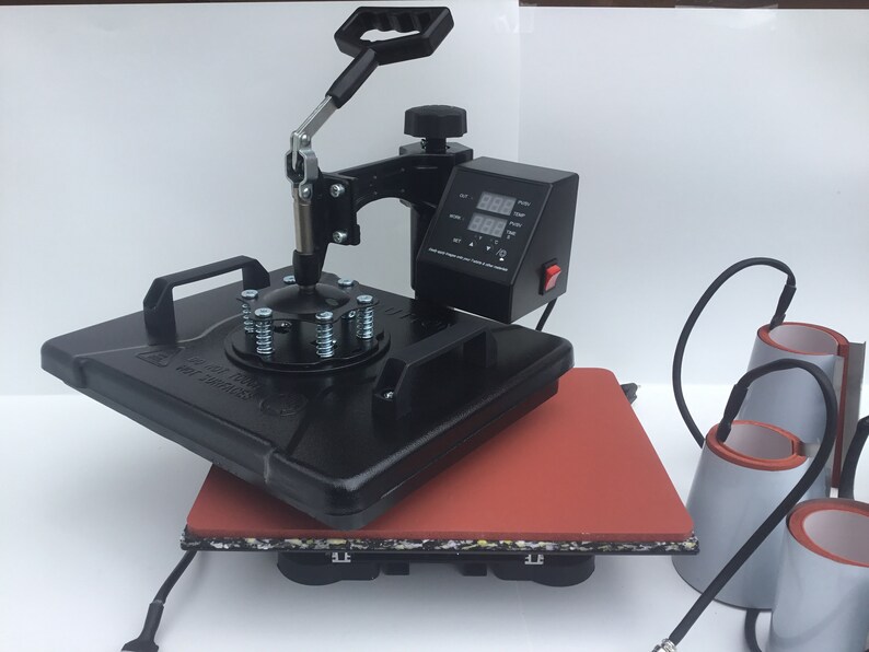 May include: Black heat press machine with a digital timer and temperature control. The machine is on a red heat-resistant mat. Three additional heat press attachments are included.