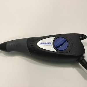May include: A black Dremel engraver with a blue speed control knob. The engraver has a silver label that reads "Dremel Engraver".