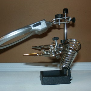May include: A silver magnifying glass with a light attached to a black stand with two adjustable clamps. The stand has a spring coil for holding small objects.