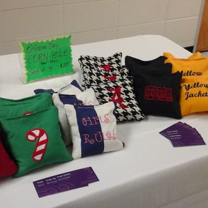 May include: A table with a variety of cornhole bags for sale. The bags are decorated with different designs, including a Christmas tree, a candy cane, a houndstooth pattern, and the words "Girls Rule". There is also a sign that says "Game Day Corn Hole Set of 4 $20".