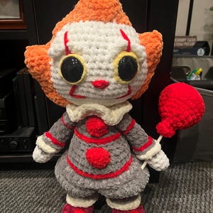 Op de afbeelding: Een gehaakt pluche speelgoed van Pennywise de dansende clown uit de film "IT". Het speelgoed draagt een grijze en rode outfit met een witte kraag en heeft een rode ballon. Het speelgoed heeft grote zwarte ogen met gele pupillen en een rode glimlach.