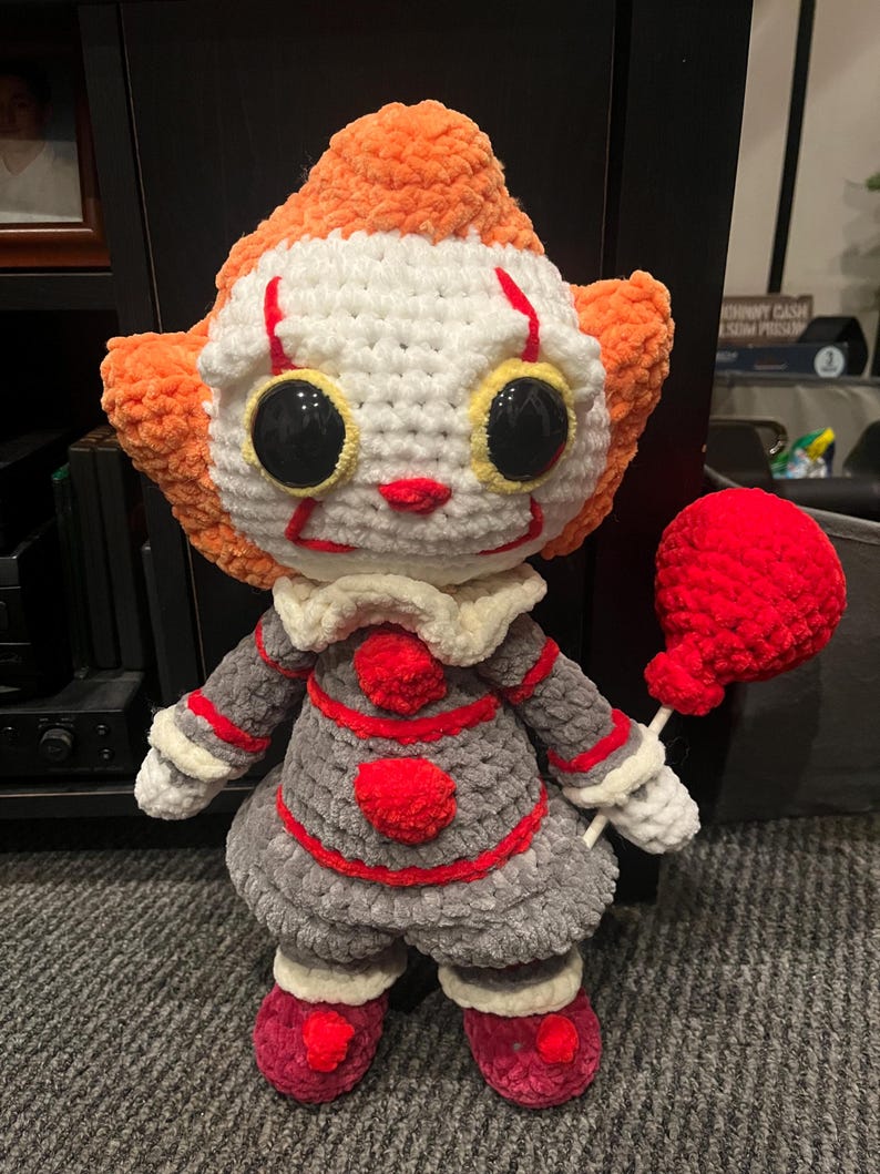 Op de afbeelding: Een gehaakt pluche speelgoed van Pennywise de dansende clown uit de film "IT". Het speelgoed draagt een grijze en rode outfit met een witte kraag en heeft een rode ballon. Het speelgoed heeft grote zwarte ogen met gele pupillen en een rode glimlach.