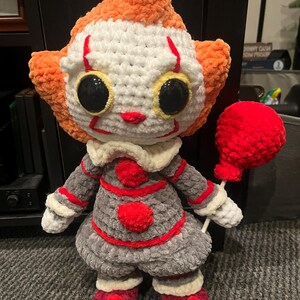 Op de afbeelding: Een gehaakt pluche speelgoed van Pennywise de dansende clown uit de film "IT". Het speelgoed draagt een rood-wit gestreepte outfit en heeft een rode ballon.