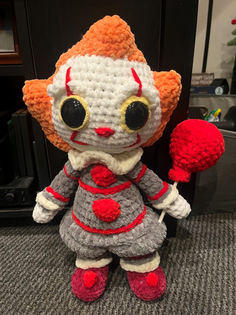Op de afbeelding: Een gehaakt pluche speelgoed van Pennywise de dansende clown uit de film "IT". Het speelgoed draagt een rood-wit gestreepte outfit en heeft een rode ballon.