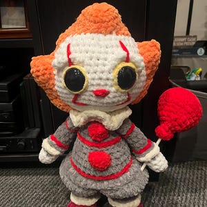 Op de afbeelding: Een gehaakt pluche speelgoed van Pennywise de dansende clown uit de film IT. Het speelgoed draagt een grijze en rode outfit en heeft een rode ballon.