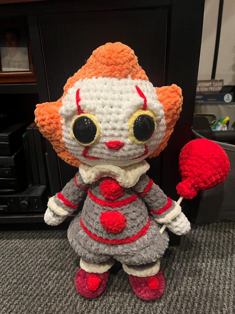 Op de afbeelding: Een gehaakt pluche speelgoed van Pennywise de dansende clown uit de film IT. Het speelgoed draagt een grijze en rode outfit en heeft een rode ballon.