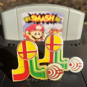 May include: Two gold-colored Super Smash Bros. pins with red, yellow, and green accents. The pins are shaped like the Super Smash Bros. logo and are attached to a Nintendo 64 game cartridge case.