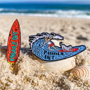 May include: Two enamel pins featuring a surfer and a dog on a surfboard with the text "Paddle Out" on a blue wave. The pins are on a sandy beach with seashells.