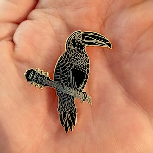 May include: Black enamel pin of a toucan perched on a guitar. The toucan has a long, curved beak and detailed feathers. The guitar is gold and has a simple design.