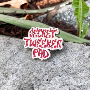 May include: A silver pin with red lettering that reads "SECRET TWEETER PAD".