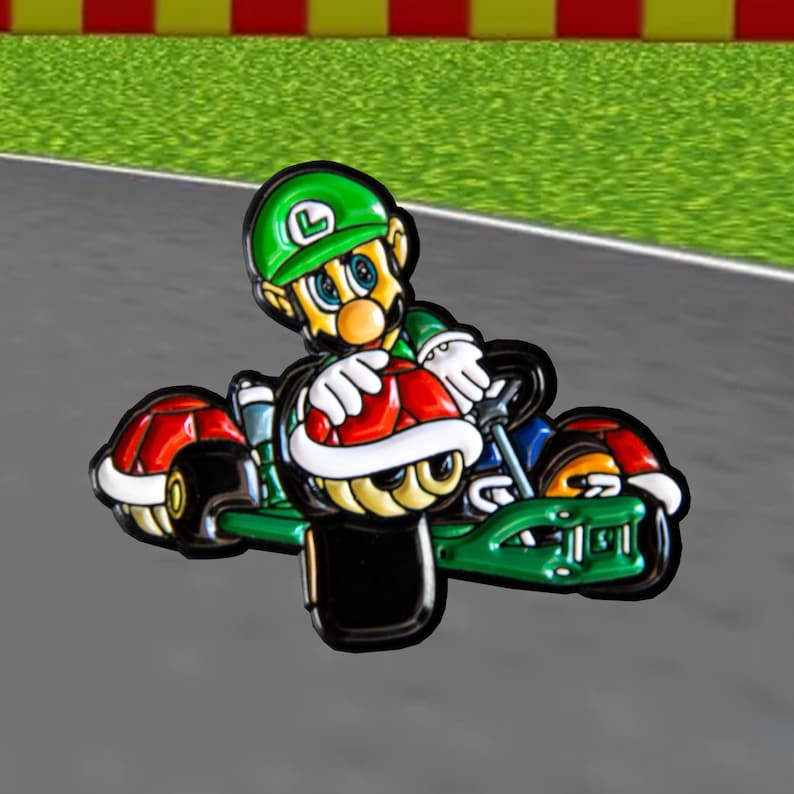 Pu&ograve; includere: Uno spillo in smalto verde e rosso di un personaggio dei cartoni animati che guida un kart. Il personaggio indossa un cappello verde con una L sopra. Il kart ha un guscio rosso e bianco sul retro.