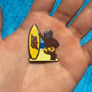May include: A yellow and red enamel pin featuring a cartoon character holding a surfboard with the word "GRIZZ" on it.
