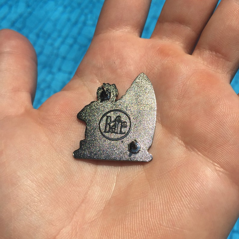 May include: A silver metal pin with a glittery finish. The pin features a cartoon surfer riding a wave. The back of the pin has the text "Baff 2020" and a small circle with the word "Baff" inside.