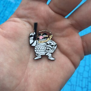 May include: A white and black enamel pin featuring a cartoon character dressed as a Stormtrooper holding a blaster. The character has brown hair, a mustache, and a pink nose.
